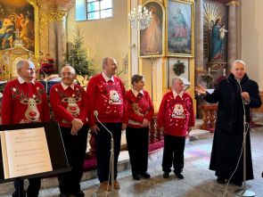 grupa mieszkańców ubranych w czerwone swetry z elementami świątecznymi z proboszczem podczas kolędowania w kościele