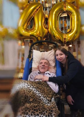 mieszkaniec w wózku terapeutycznym pozuje do zdjęcia, obok niego terapeutka a w tle dmuchana balonowa liczba 40 