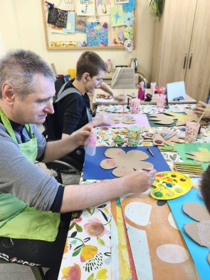 dwoje mieszkańów w fartuchach wykonuje prace plastyczne przy długim stole z róznymi materiałami plastycznymi