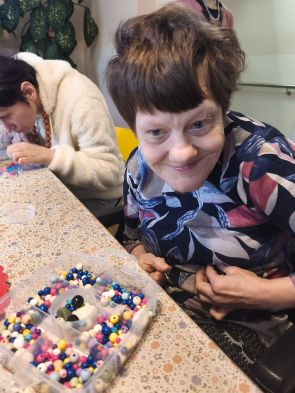 mieszkanka podczas zajęć terapeutycznych, na długim stole koraliki, w tle kolejna mieszkanka z kolorowymi koralami