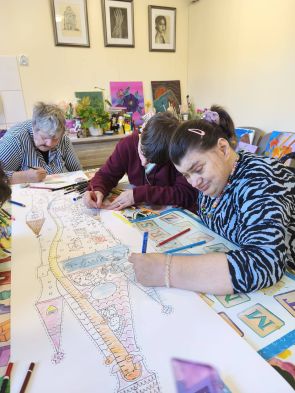 troje mieszkanek kredkami wypełnia wspólną prace na wielkim stole, w tle zaramowane prace plastyczne