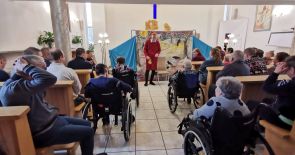 Grupa mieszkańców siędzących w ławkach lub w wózkach inwalidzkich w kaplicy domu podczas przedstawienia teatralnego. Przed ołtarzem zaimprowizowane kolorowe tło sceniczne a przed nim aktor.
