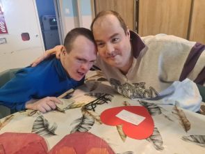 Dwóch mieszkańców podczas zajęć terapeutycznych pozuje do zdjęcia obejmując się. Na stole widać przygotowywaną kartę walentynkową w kształcie serca w kolorze czerwonym.