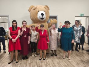 Kilka mieszkanek domu odświętnie ubranych pozuje do zdjęcia z 3 metrowym pluszowym misiem. Osoby ustawione są w holu o jasnych ścianach, za nimi widać drzwi do kolejnych pomieszczeń.